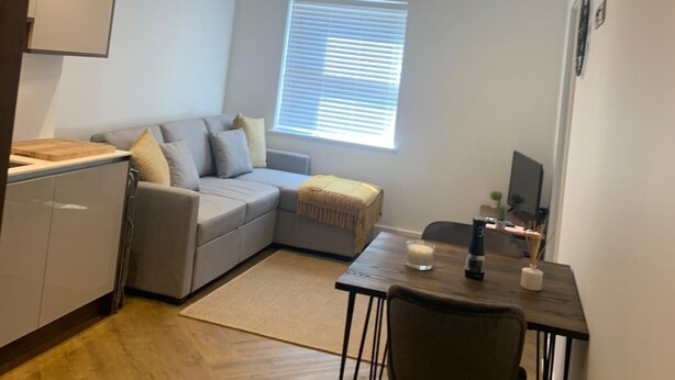 A compact living space with a kitchenette, a grey sofa, a dining table with two chairs, and a window with blinds. - Unique 1 bedroom in Reading (Reading)