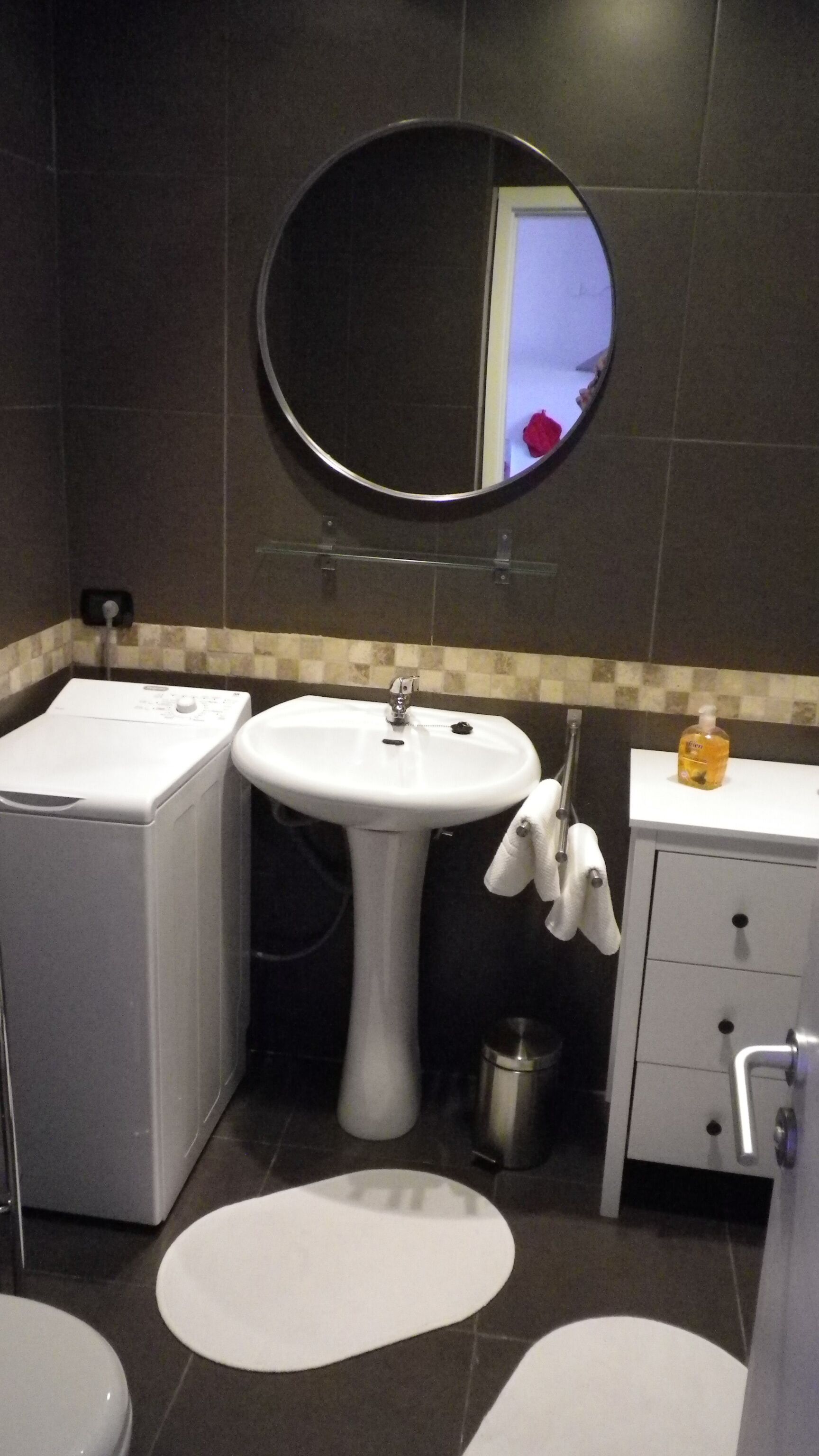 A compact bathroom with a pedestal sink, a round mirror, a white toilet, and a small cabinet with a bottle of hand soap.