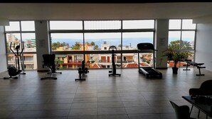 Un gimnasio con equipo de ejercicio y ventanas amplias que ofrecen vistas a edificios y al mar.