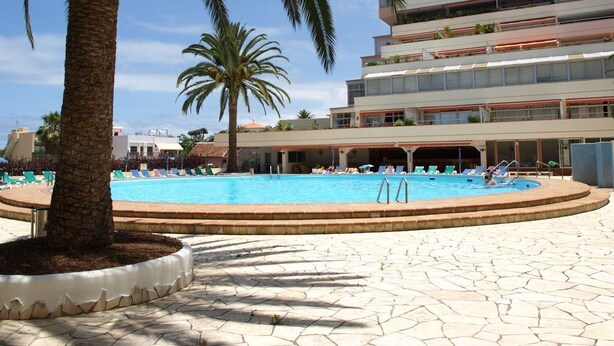 Un área de piscina con sillones reclinables y palmeras, rodeada por un edificio de varios pisos con balcones.