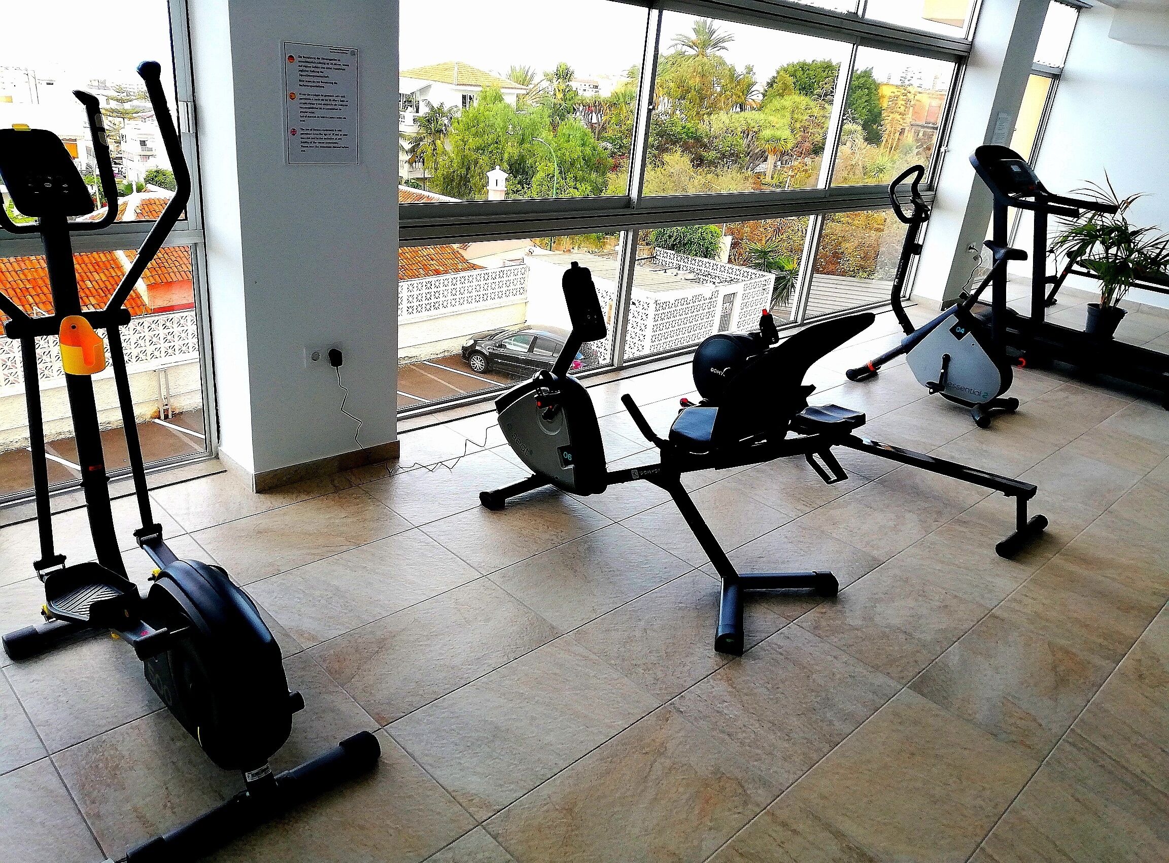 Un gimnasio con bicicletas estáticas y equipo cardiovascular, ventanas amplias con vistas a edificios y árboles, y suelo de baldosas.