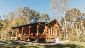A log cabin with a porch, stone foundation, and a green roof, surrounded by trees. - Old World Craftmanship with Modern Amenities (Bristol)