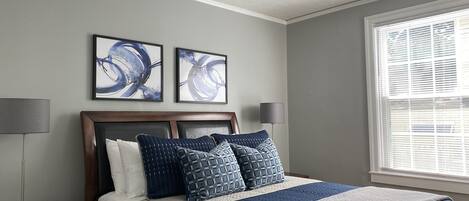 A bedroom with a bed, two wall-mounted abstract paintings, a ceiling fan, a bedside lamp, and a window with blinds.