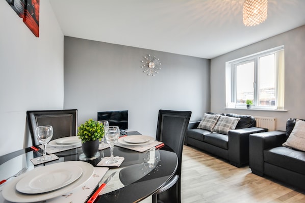 A modern living room with a dining area. There is a dining table set with plates, glasses, and cutlery. A black sofa and a black chair are visible in the living area.