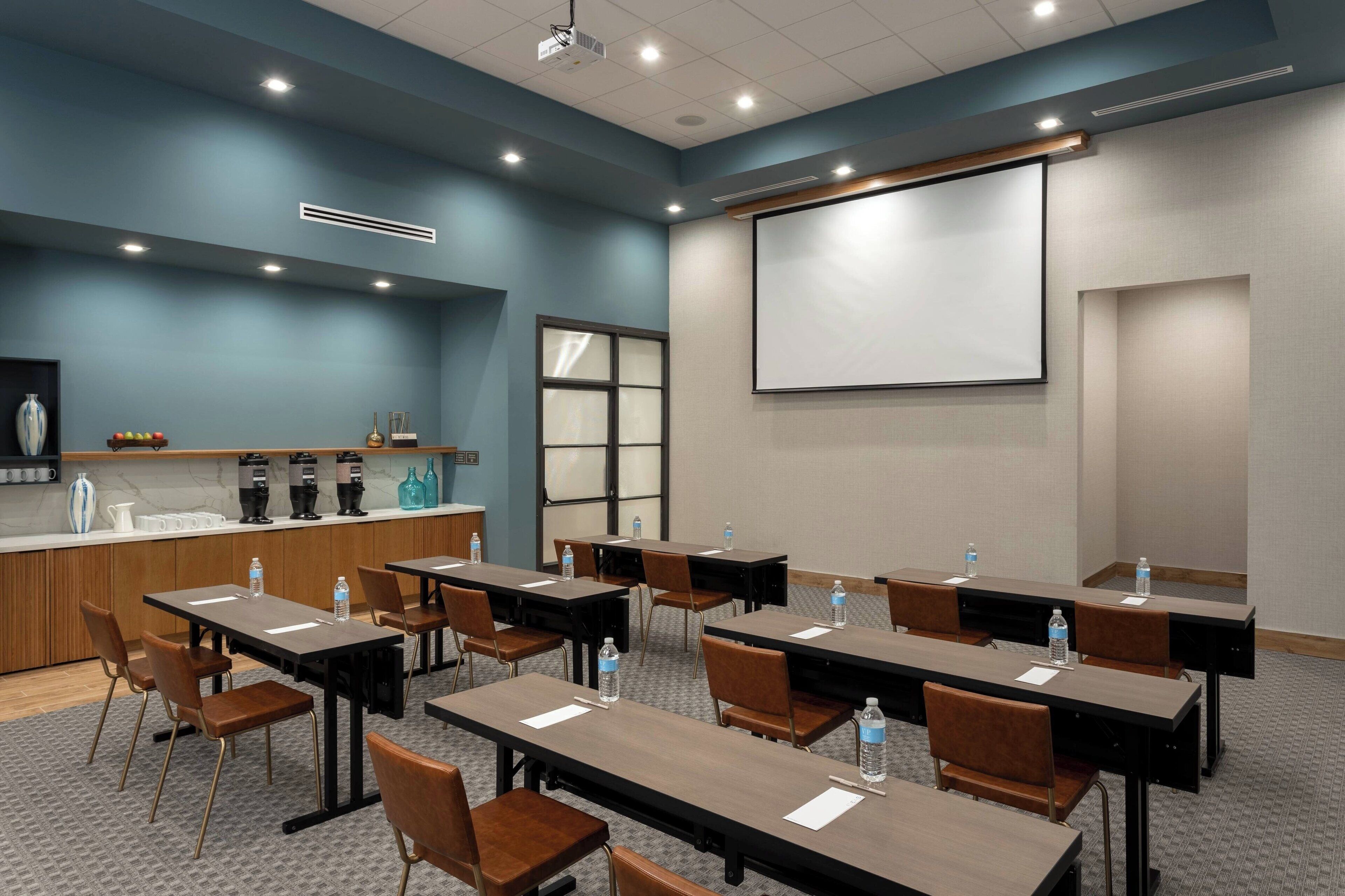 Uma sala de conferências com mesas, cadeiras, tela de projetor e um espaço para café.