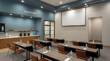 Uma sala de conferências com mesas, cadeiras, tela de projetor e um espaço para café.