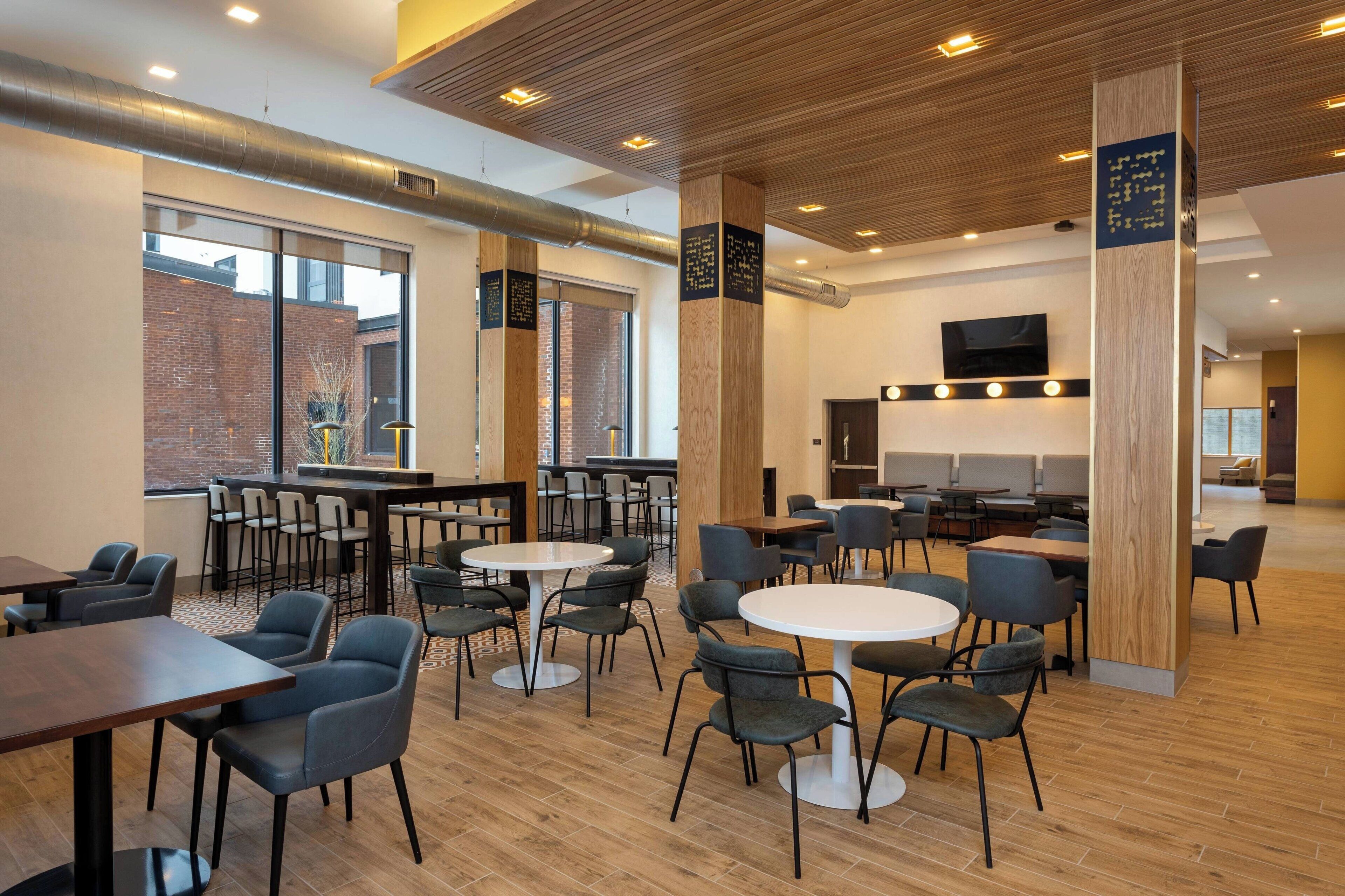 A modern dining area with round tables and chairs, wooden accents, and a brick wall visible through large windows.