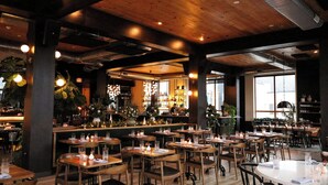 Een restaurant met houten tafels en stoelen, een bar en grote ramen voor natuurlijk licht.