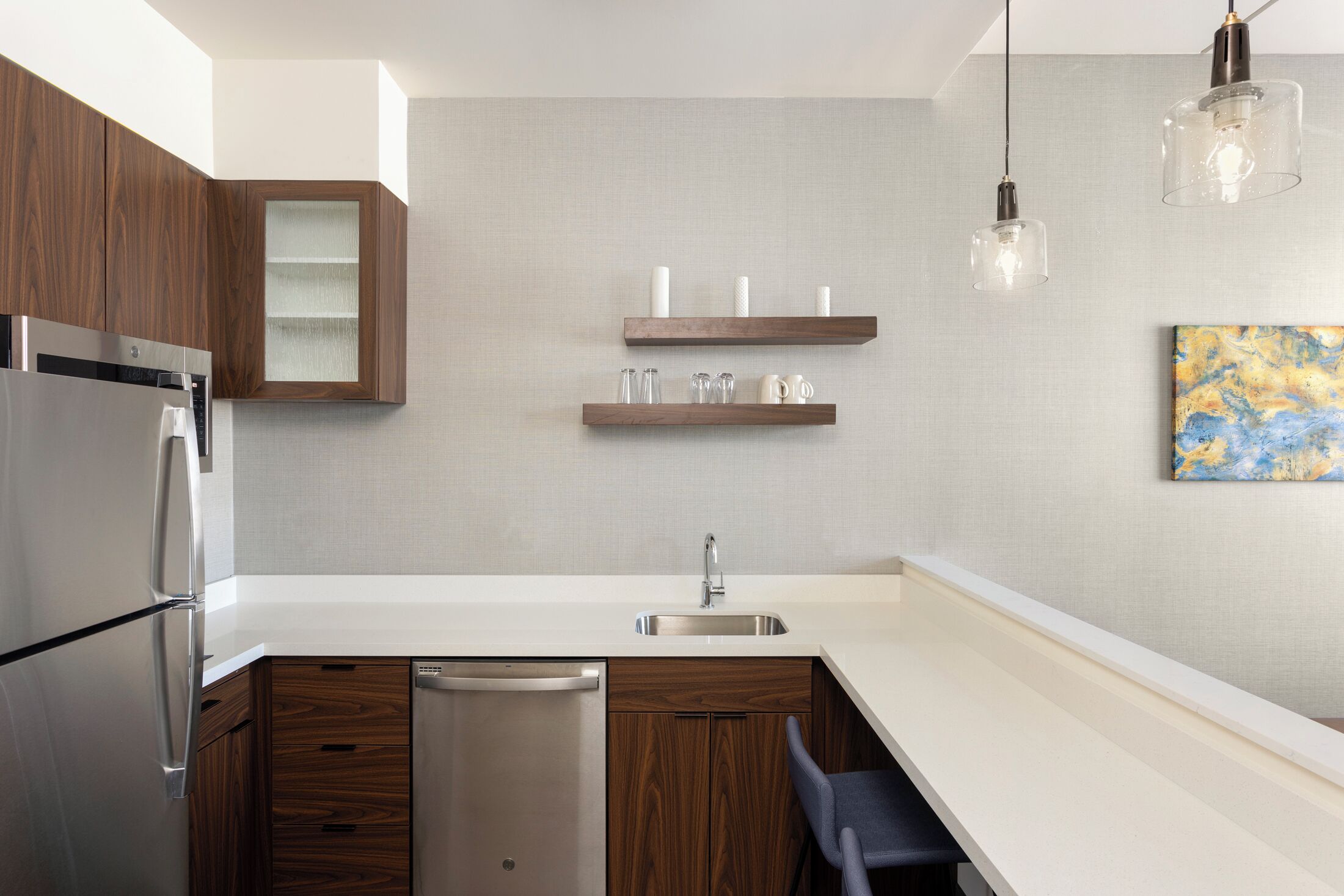 A modern kitchen with wooden cabinets, a stainless steel refrigerator, a dishwasher, and a white countertop. There are two hanging pendant lights and a painting on the wall.