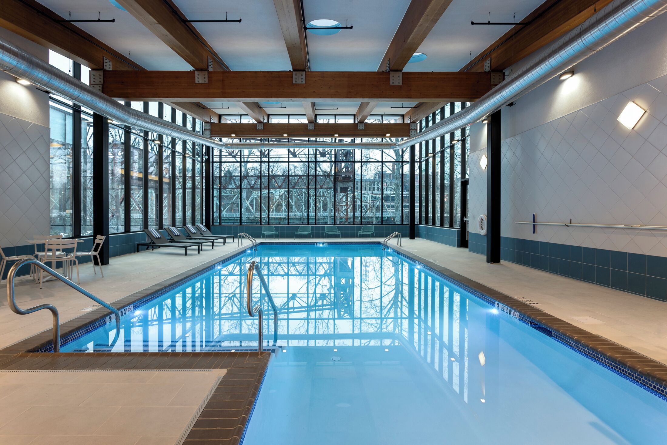An indoor swimming pool with clear glass walls, wooden beams, and a tiled floor.
