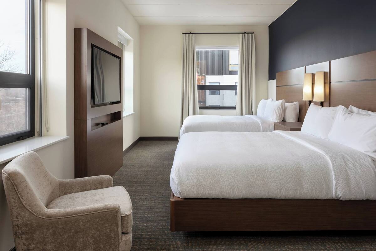 A hotel room with a large bed, a TV mounted on the wall, a chair, and a window with curtains.