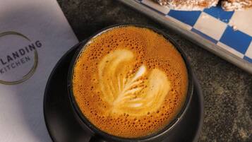 Una taza de café con arte de latte sobre un platito negro, un pastel sobre una bandeja a cuadros y una servilleta con un logotipo.
