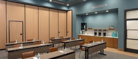 A conference room with tables, chairs, a kitchenette, and a large screen.