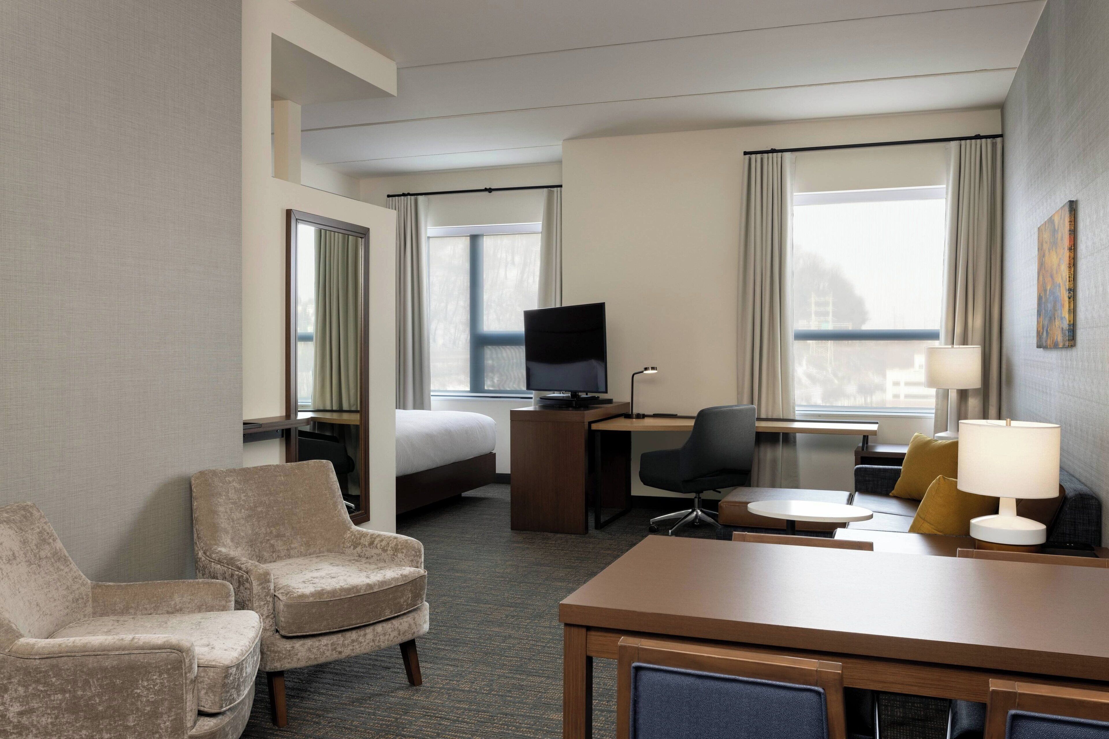 A hotel room with a bed, a desk with a computer, two armchairs, a small table, and a lamp.