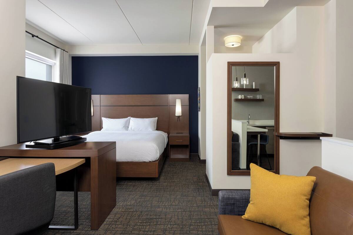 A hotel room with a bed, a desk with a TV, a sofa, a mirror, and a small table with a lamp.