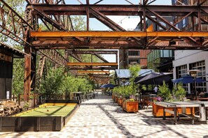 An outdoor dining area with tables, chairs, and umbrellas, set against a backdrop of a large metal structure and greenery. - Residence Inn by Marriott Philadelphia Bala Cynwyd (Bala-Cynwyd)
