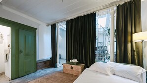 Una camera d'albergo con un letto, una porta verde, una finestra con tende e un piccolo tavolino con un cesto.