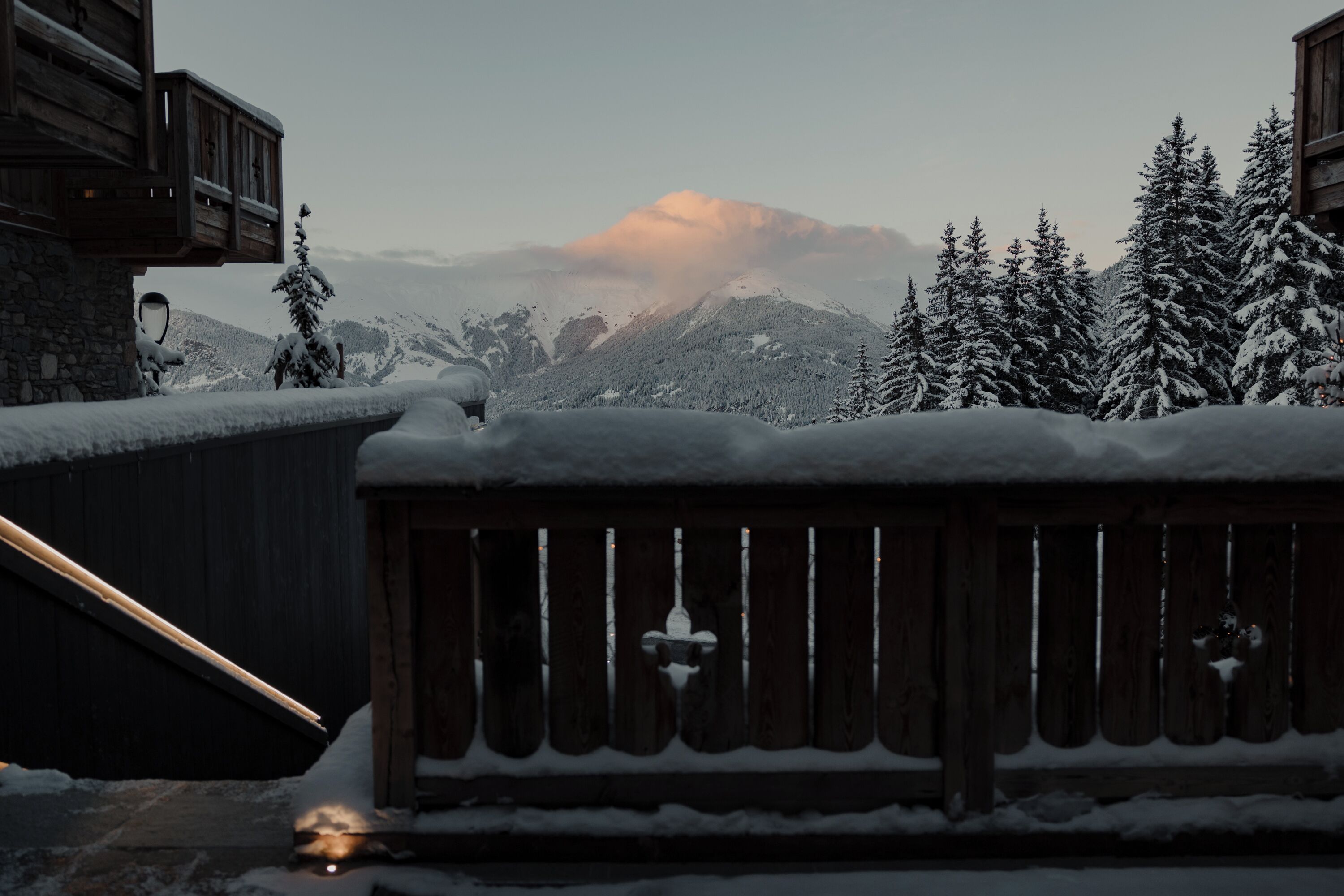 A wooden balcony with snow-covered railings, a stone wall, and a view of snow-laden trees and mountains in the background.