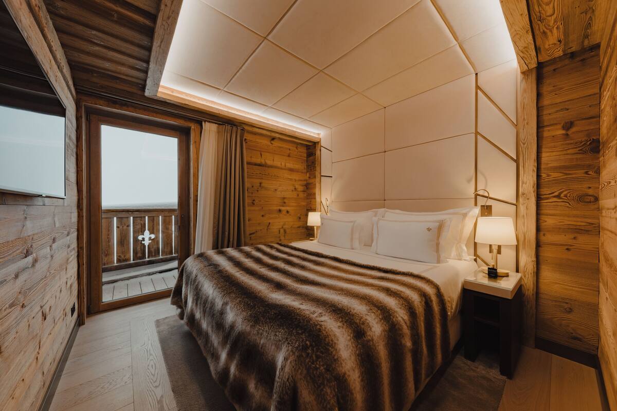 Quarto estilo cabana de madeira com uma cama grande, uma mesinha de cabeceira e uma janela com vista para uma varanda.