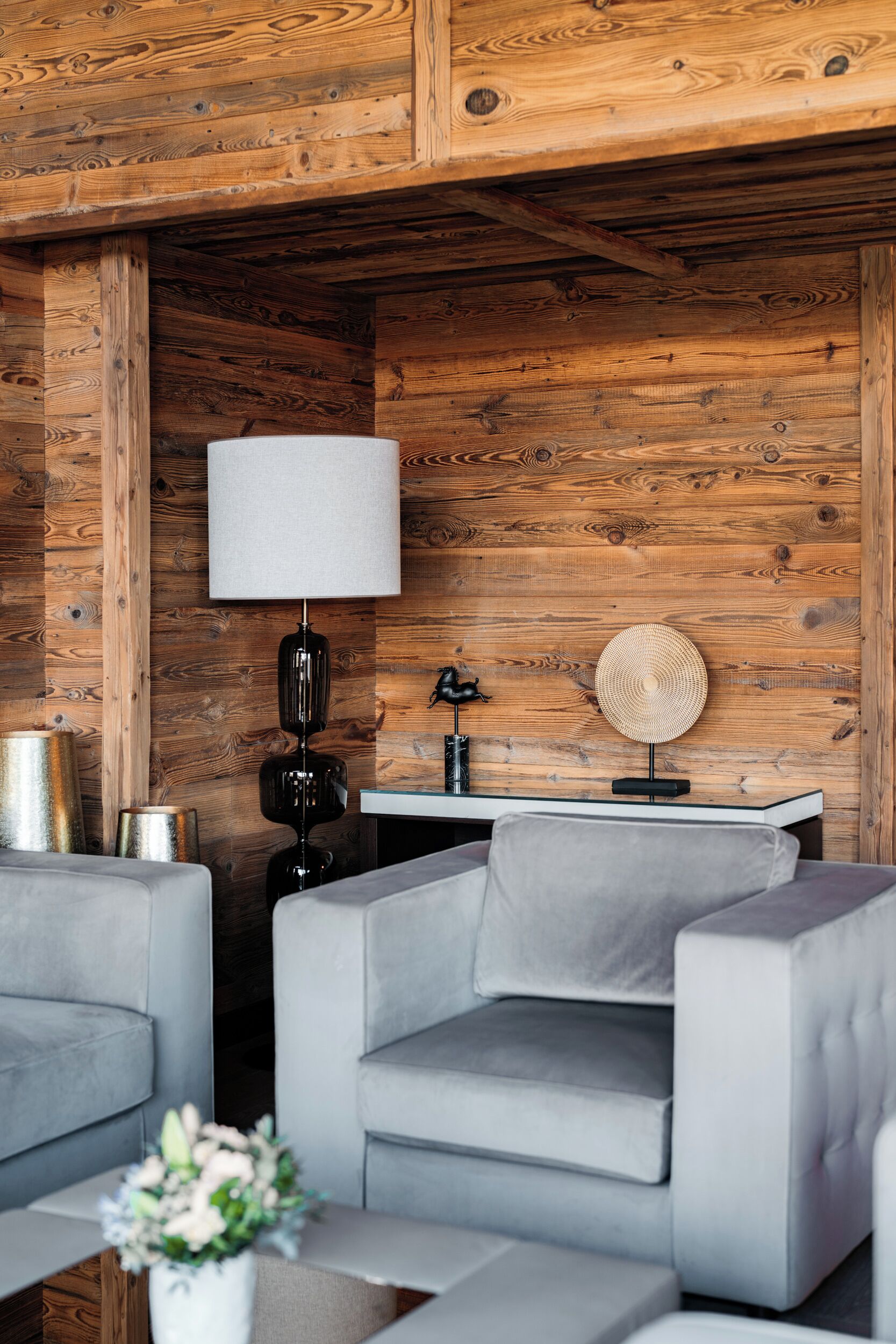 A modern living room with a wooden wall, a grey sofa, a round table, a lamp, and a vase with flowers.
