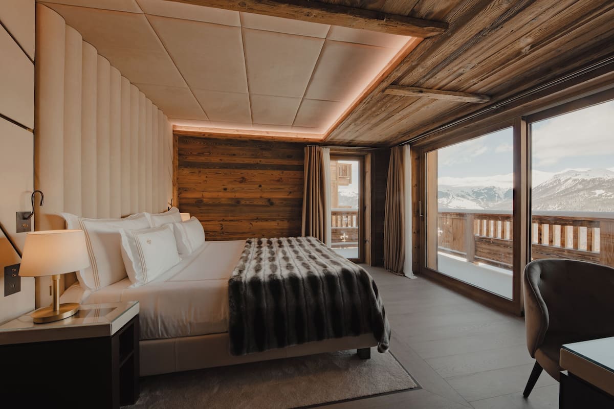 Quarto com uma cama grande, mesa de cabeceira, cadeira e vista para montanhas cobertas de neve.