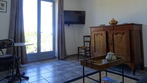 Ein Zimmer mit einem Holzkasten, einem an der Wand befestigten Fernseher, einem Esstisch mit Stühlen und einem großen Fenster mit Vorhängen.