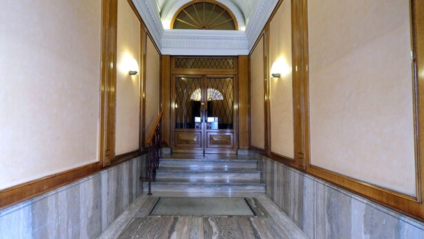 Un couloir au sol de marbre, avec une porte en bois au bout, et de part et d’autre, des appliques murales.