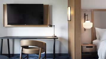 Ein Hotelzimmer mit flachbildfernseher an der Wand, einem Schreibtisch mit Stuhl, einem Nachttisch mit Lampe und einem Bett mit weißer Bettwäsche.