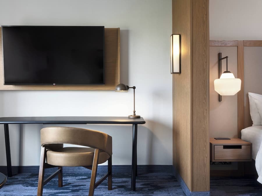 Ein Hotelzimmer mit flachbildfernseher an der Wand, einem Schreibtisch mit Stuhl, einem Nachttisch mit Lampe und einem Bett mit weißer Bettwäsche.