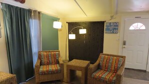 Une pièce comprenant deux fauteuils en osier, une petite table, un tapis à motifs, un rideau vert et un mur orné d’une enseigne.