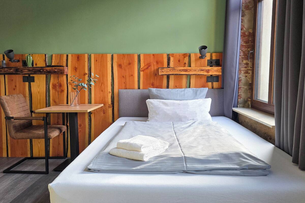 A single bed with a white comforter and pillows, a wooden chair, a small table with a vase, and a shelf with decorative items.