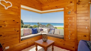 A cozy room with a wooden interior, a large window offering a view of the beach and ocean, a cushioned seating area with pillows, and a small table with a decorative item.