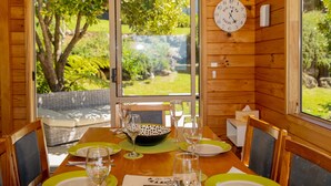 A wooden cabin dining area with a table set for four, a clock on the wall, and a view of greenery outside.