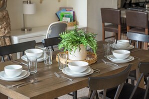 Une table à manger dressée avec de l’assiette blanche, des bols, des verres et des couverts, agrémentée d’une plante en pot au centre.