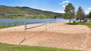 A sandy volleyball court with a net, surrounded by grass and trees, near a lake and hills.