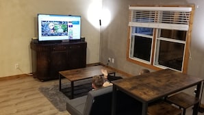 A room with a wooden table, a television on a stand, a window with blinds, and a grey sofa. - Clean and Cozy Creekside Home (Conroe)