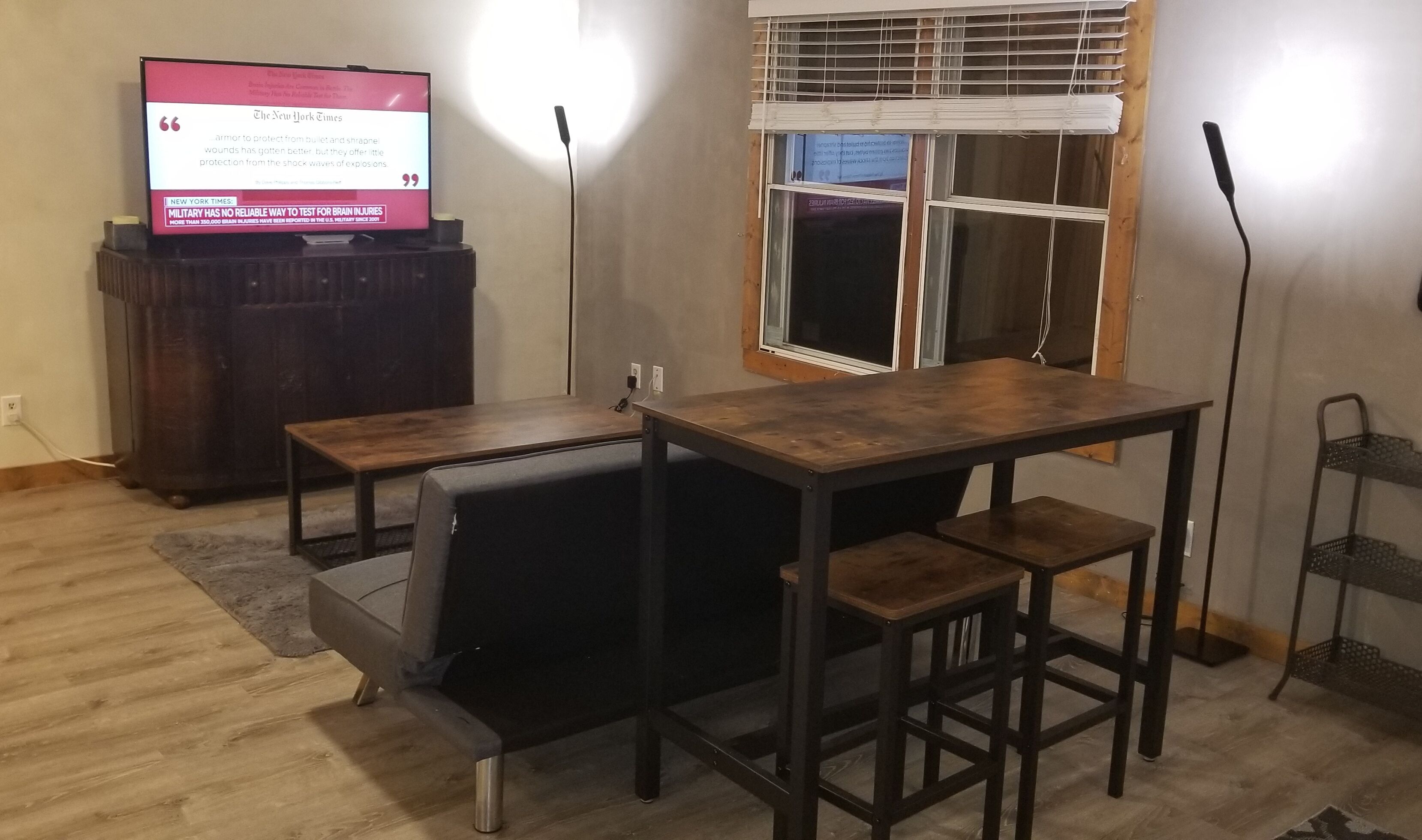 A small, furnished room with a wooden table and bench, a television on a stand, and a window with blinds.