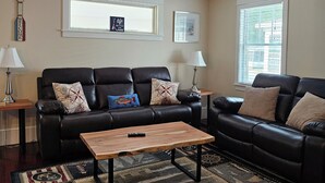 A living room with a black leather sofa, a wooden coffee table, and a patterned rug. There are two windows with blinds, a wall-mounted lamp, and a framed picture. - Newly updated,close to downtown and the lake, pet friendly  (Grand Haven)