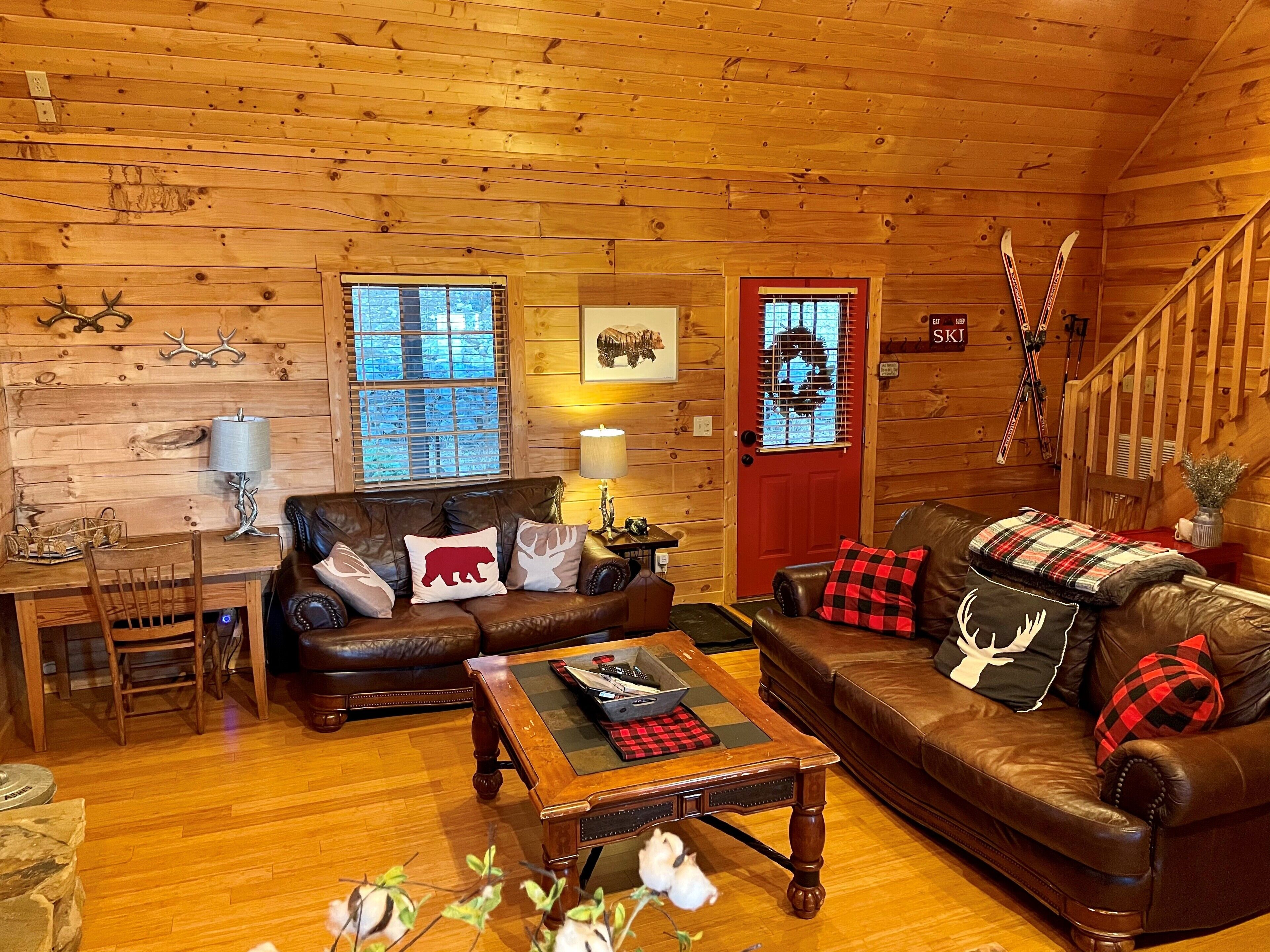 A cozy log cabin interior with a wooden floor, leather sofas, a coffee table, and a staircase.