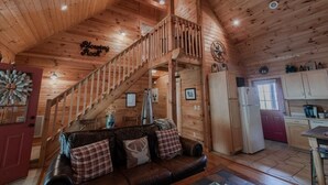 Interior de una cabaña de madera con una escalera que sube a un altillo, una sala de estar con un sofá de cuero y una zona de cocina.
