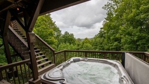 Un jacuzzi en una terraza de madera con una escalera que lleva a una zona cubierta, rodeado de abundante vegetación.