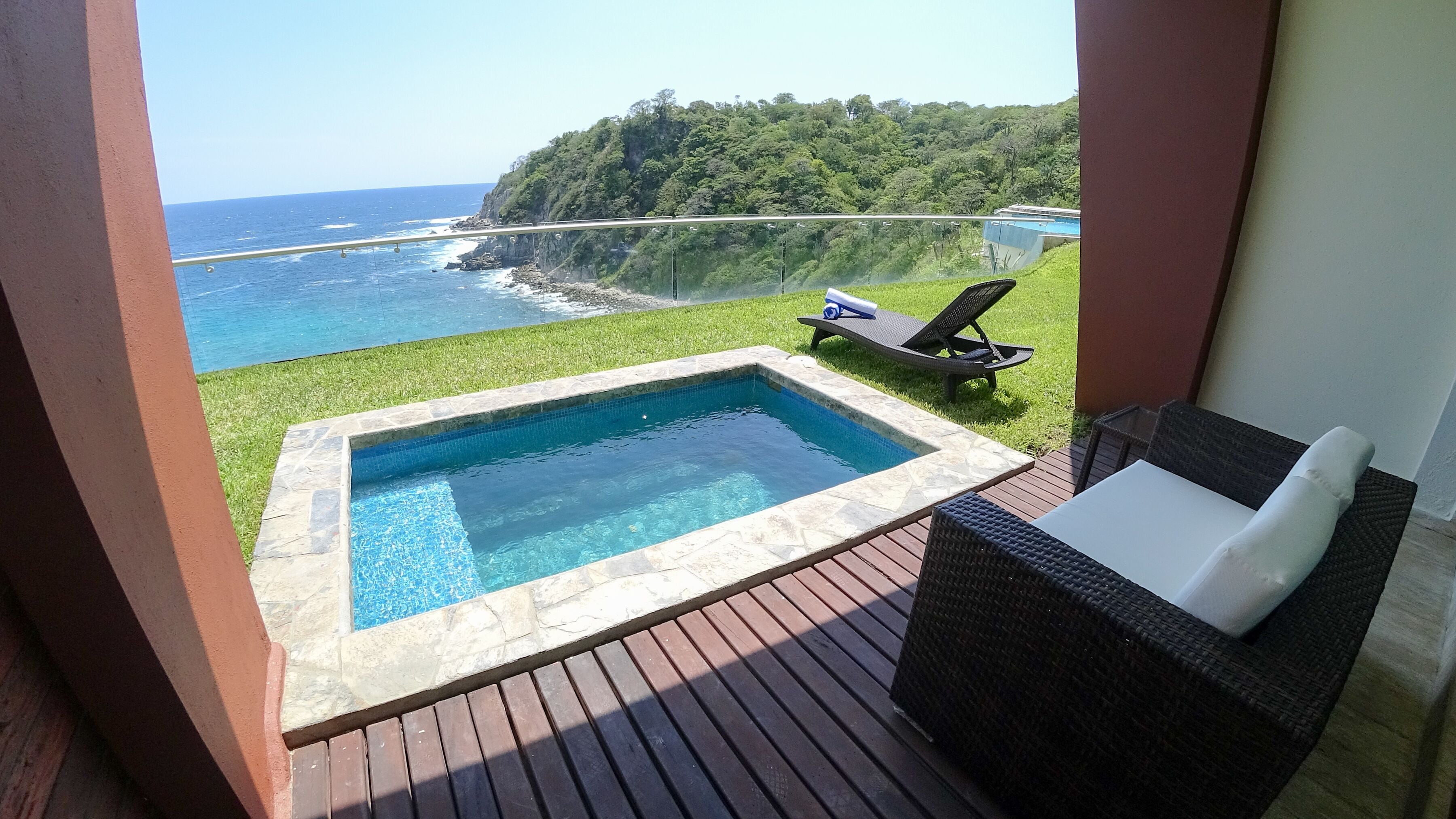 Un balcón con piscina, una silla de descanso y vistas al océano y una colina boscosa.