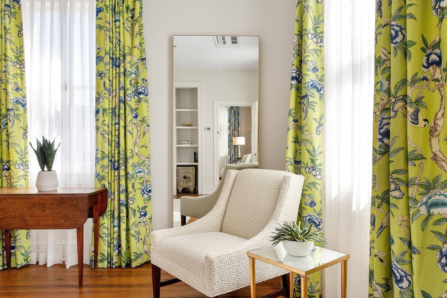 Een kamer met een houten eettafel, een gestreept fauteuil, een klein bijzettafeltje met een plant en gele bloemen gordijnen.