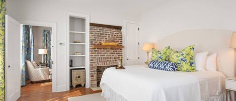 A bedroom with a white bed, a brick fireplace, and a wooden floor.