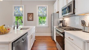 Una cocina moderna con armarios blancos, lavavajillas de acero inoxidable, horno empotrado y microondas. Hay dos ventanas con persianas decorativas, un fregadero con grifo y una encimera con una cesta de fruta.
