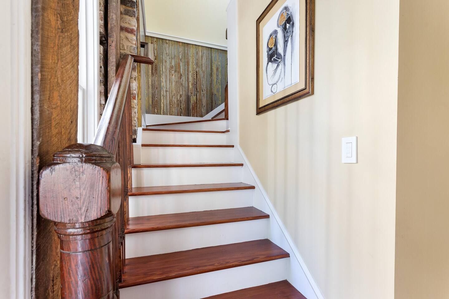 Una escalera de madera con escalones blancos y barandilla de madera, que conduce a una habitación con puerta de madera y un cuadro enmarcado en la pared.