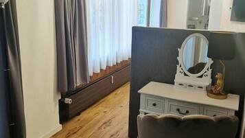 Ett rum med trägolv, en vit byrå med lådor, en spegel och en liten staty. Det finns ett fönster med gardiner, och en element syns på väggen.