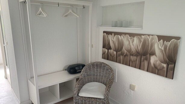 Una habitació amb un armari blanc, una cadira de vimet, un aparell de ràdio i una estanteta penjada a la paret amb un quadre floral.