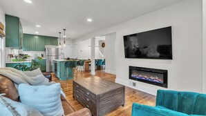 Un salon moderne avec un canapé en cuir marron, des fauteuils turquoise et une table basse en bois. Un téléviseur à écran plat et une cheminée sont fixés au mur. La cuisine, visible à l’arrière-plan, est dotée de meubles verts et d’appareils électroménagers en acier inoxydable.