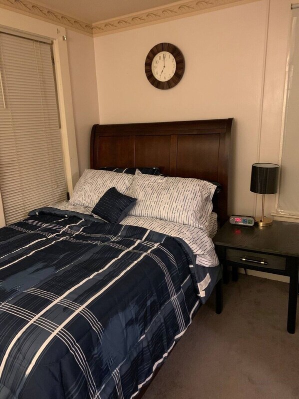 A bedroom with a dark wood headboard, a bed with striped bedding, a bedside table with a clock, and a window with blinds.